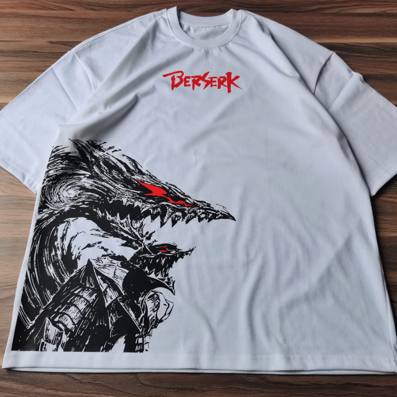 تيشيرت أبيض أوفرسايز عليه تصميم لشخصية جاتس من أنمي بيرزيرك بداخل البيرزيركر آرمور باللون الأسود مع تفاصيل حمراء من متصور من تحت مقدم من بخلفية خشبية Sugoi Store