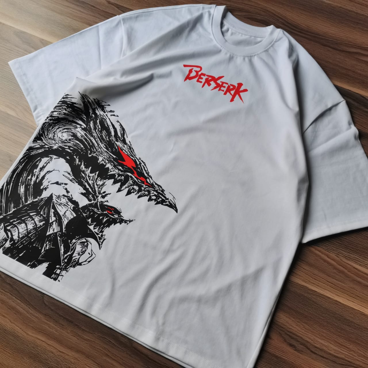 تيشيرت أبيض أوفرسايز عليه تصميم لشخصية جاتس من أنمي بيرزيرك بداخل البيرزيركر آرمور باللون الأسود مع تفاصيل حمراء من متصور من الجانب اليمين مقدم من بخلفية خشبية Sugoi Store