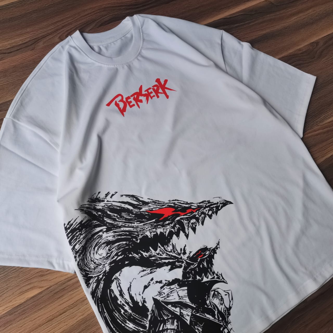 تيشيرت أبيض أوفرسايز عليه تصميم لشخصية جاتس من أنمي بيرزيرك بداخل البيرزيركر آرمور باللون الأسود مع تفاصيل حمراء من متصور من الجانب الشمال مقدم من بخلفية خشبية Sugoi Store